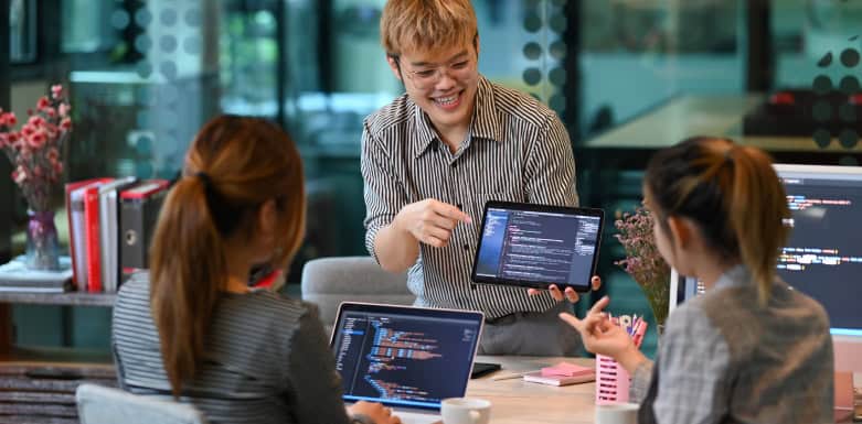 HTML5: evolução, funcionalidades e boas práticas Mulher loira mostrando código de HTML no tablet para outras duas mulheres, que estão sentadas no escritório de trabalho.
