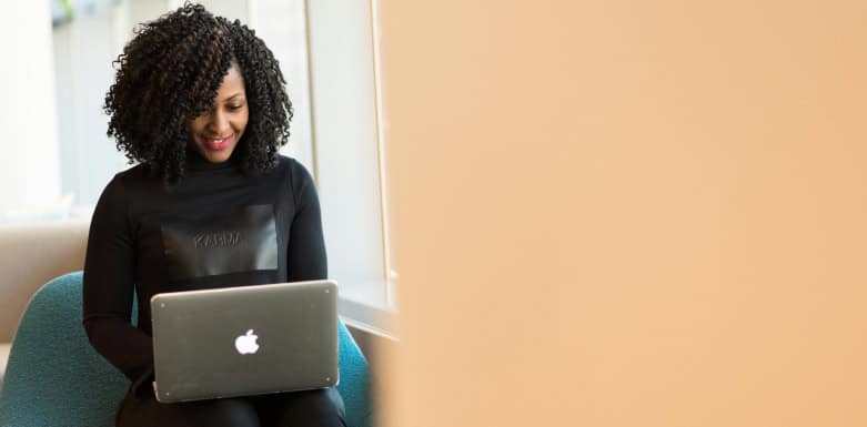O que é SEO programático? Mulher de roupa preta, sentada numa poltrona azul, enquanto digita num notebook prata, que está em seu colo.