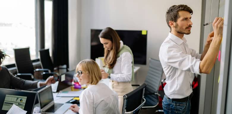 Atributo Href: saiba como afeta o seu projeto de SEO! Três colaboradores numa sala de reunião. O homem escreve no quadro e as mulheres estão olhando no computador.