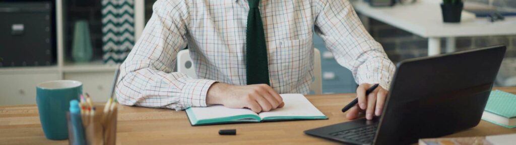 Homem de camisa xadrez e gravata preta, de frente para o notebook, numa mesa de madeira clara. Uma de suas mãos está sobre um caderno e a outra no teclado do notebook.