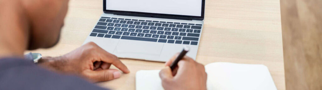 Homem anotando em bloco de notas, em frente ao notebook.